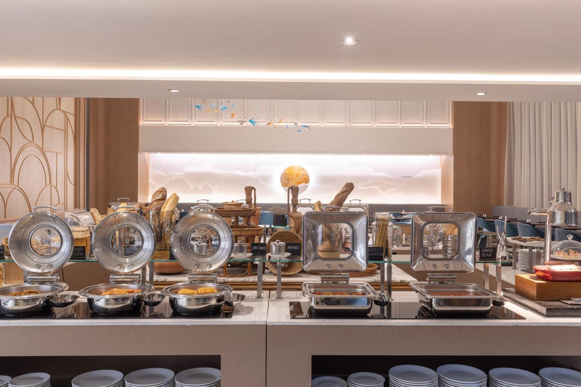 Siam Mandarina Hotel - Free Bkk Airport Shuttle Samut Prakan Exterior photo The photo shows a modern buffet setup in a dining area. There are multiple food warmers with transparent lids positioned neatly on a countertop. Behind them, there are various food items displayed, including what appears to be bread, possibly in a de