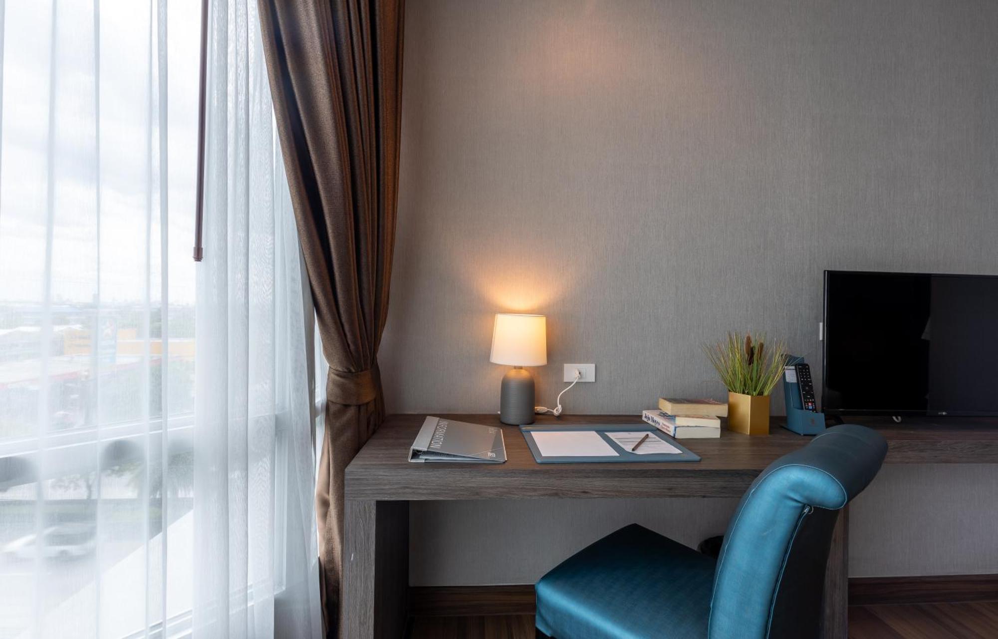 Siam Mandarina Hotel - Free Bkk Airport Shuttle Samut Prakan Exterior photo The photo showcases a modern workspace set up in a room. There is a wooden desk with a laptop and some stationery items on it, including papers and a notebook. A small lamp provides warm light, and a green plant adds a touch of decor. The chair is up